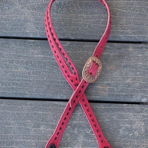 May include: A red leather guitar strap with black stitching and a decorative metal buckle. The strap is designed with a diamond pattern and has a braided detail at the ends.