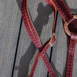 May include: Close-up of a burgundy leather horse bridle with pink stitching and accents. The bridle features a silver buckle and decorative metal hardware. The text "Windy Wyo Leather Co" is visible in the upper left corner.