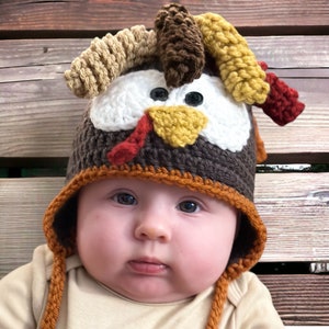 May include: A baby wearing a brown, yellow, and red crocheted turkey hat with a white face and black eyes.