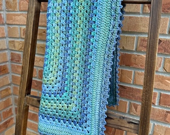 May include: A crocheted baby blanket in shades of blue and green, with a scalloped border. The blanket is hanging on a wooden ladder.