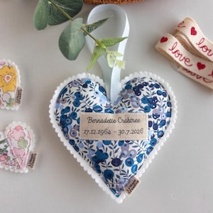 May include: A heart-shaped fabric ornament featuring a blue and white floral pattern with a white pom-pom trim edge. A beige fabric label is sewn to the centre with the text "Bernadette Crabtree" and "17.12.1964 - 30.7.2026" in black ink.