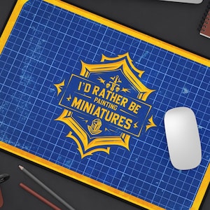 May include: A rectangular blue and yellow mousepad with the text "I'D RATHER BE PAINTING MINIATURES." A white mouse sits on the pad. The pad has a grid pattern and a yellow border. A laptop, pencils, and a coffee cup are also visible.