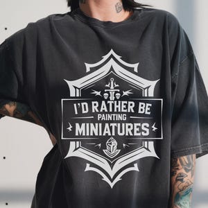 May include: Dark gray t-shirt with a white graphic that reads "I'D RATHER BE PAINTING MINIATURES." The graphic is framed by a decorative shield design with a sword, skull, and helmet. The shirt has short sleeves and a relaxed fit.