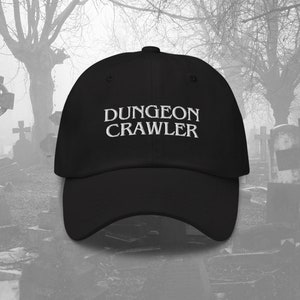 May include: Black baseball cap with white embroidered text that reads "DUNGEON CRAWLER". The cap is photographed in front of a foggy graveyard.
