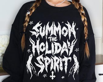 Op de afbeelding: Zwarte sweatshirt met de witte tekst "SUMMON THE HOLIDAY SPIRIT" in gotische letters. Het ontwerp bevat sterren, een kruis en gestileerde handen. De sweatshirt heeft een ronde hals en lange mouwen.