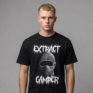 Puede incluir: Camiseta negra con las palabras "EXTRACT CAMPER" en blanco, con una fuente gótica estilizada. La camiseta presenta un gráfico de un pasamontañas. El modelo lleva vaqueros azules.