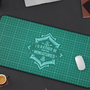 May include: A rectangular green cutting mat with a grid pattern and the text "I'D RATHER BE PAINTING MINIATURES" in a teal emblem. A white computer mouse is on the right side.