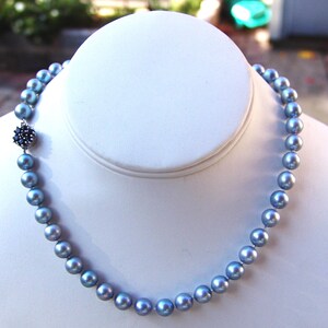 May include: A blue-grey pearl necklace displayed on a white bust. The necklace features round pearls and a decorative clasp with dark blue accents. The pearls have a subtle luster, and the clasp adds a touch of elegance to the design.