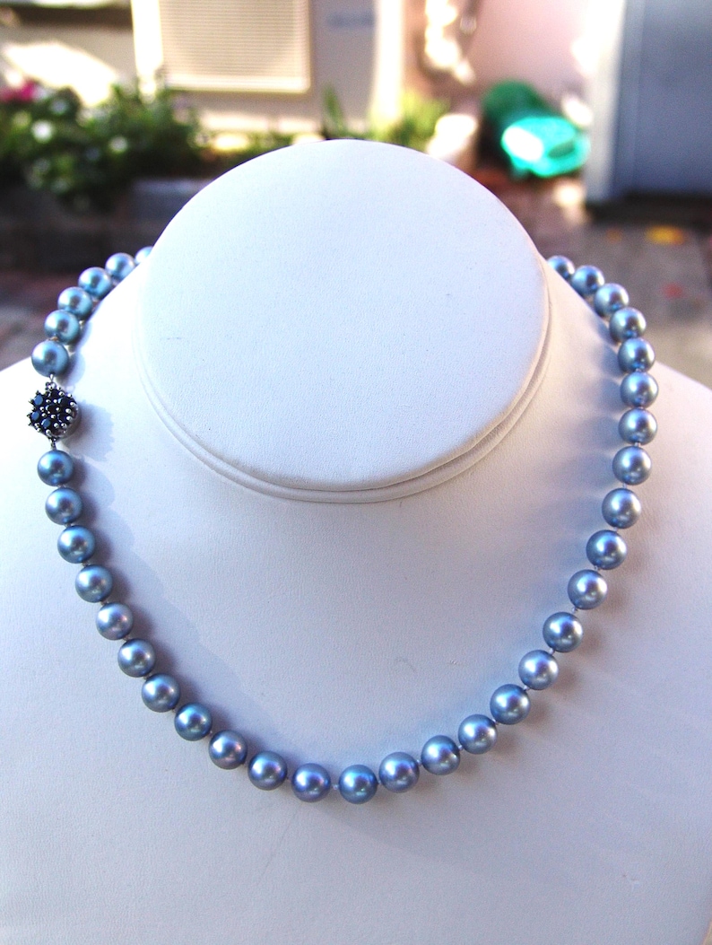 May include: A blue-grey pearl necklace displayed on a white bust. The necklace features round pearls and a decorative clasp with dark blue accents. The pearls have a subtle luster, and the clasp adds a touch of elegance to the design.