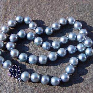 May include: A necklace composed of light blue pearls, arranged in a circular pattern. The pearls have a lustrous sheen and are connected by a delicate string. The clasp is a dark blue floral design.