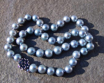 Op de afbeelding: Een ketting bestaande uit lichtblauwe parels, gerangschikt in een cirkelvormig patroon. De parels hebben een glanzende glans en zijn verbonden door een delicate draad. De sluiting is een donkerblauw bloemmotief.