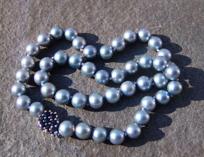 May include: A necklace composed of light blue pearls, arranged in a circular pattern. The pearls have a lustrous sheen and are connected by a delicate string. The clasp is a dark blue floral design.