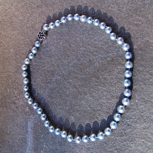 May include: A necklace featuring a strand of gray pearls. The pearls are round and evenly sized, strung together with a dark cord. A small, silver-toned clasp with a dark blue stone is visible at the closure. The necklace is displayed on a textured, gray surface.