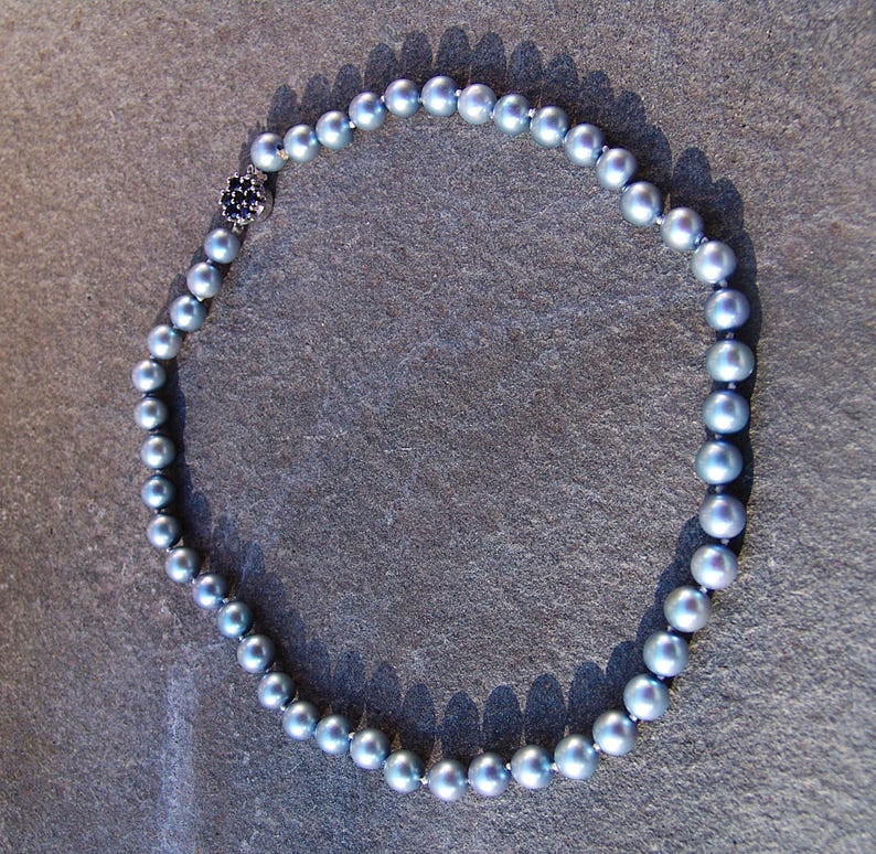 May include: A necklace featuring a strand of gray pearls. The pearls are round and evenly sized, strung together with a dark cord. A small, silver-toned clasp with a dark blue stone is visible at the closure. The necklace is displayed on a textured, gray surface.