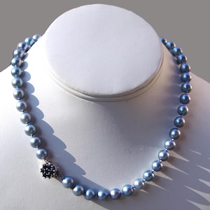 May include: A necklace of iridescent blue-gray pearls. The pearls are strung together and fastened with a dark blue floral clasp. The necklace is displayed on a white bust form against a gray background.