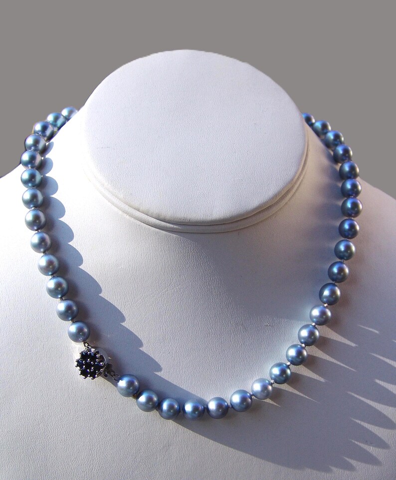 May include: A necklace of iridescent blue-gray pearls. The pearls are strung together and fastened with a dark blue floral clasp. The necklace is displayed on a white bust form against a gray background.