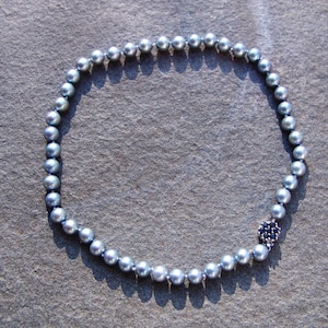 May include: A necklace featuring a strand of gray pearls. The necklace has a dark blue floral clasp with small, dark blue stones. The pearls are round and evenly spaced, creating a classic and elegant look.