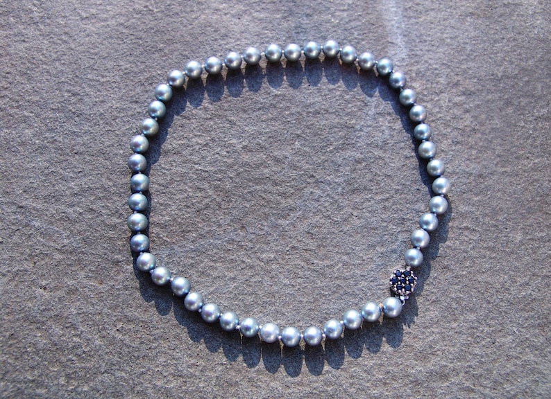 May include: A necklace featuring a strand of gray pearls. The necklace has a dark blue floral clasp with small, dark blue stones. The pearls are round and evenly spaced, creating a classic and elegant look.