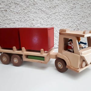 May include: A wooden toy truck with two red cargo boxes. The truck has a driver and passenger in the cab. The truck is made of natural wood and has a green base.