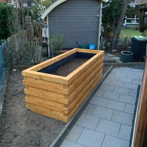 Può includere: Un letto rialzato da giardino in legno con una fodera in tessuto da paesaggio nero. Il letto è fatto di assi di legno naturale ed è situato accanto a un capanno grigio e un vialetto pavimentato.