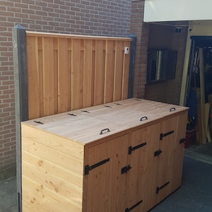 Könnte beinhalten: Ein hölzerner Außenlagerbehälter mit drei Fächern und Klapptüren. Der Behälter ist aus hellbraunem Holz mit schwarzen Metallscharnieren und Griffen. Der Behälter steht auf einer schwarzen Terrasse.