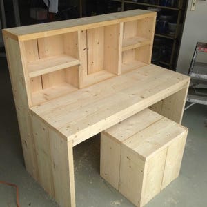 Puede incluir: Un escritorio de madera con una estantería incorporada. El escritorio tiene un acabado de madera clara y está hecho de tablones de madera sin terminar. La estantería tiene tres estantes y está abierta en la parte delantera. El escritorio está sobre un suelo de hormigón.