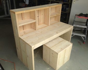 Op de afbeelding: Een houten bureau met een ingebouwde plank. Het bureau heeft een lichte houtkleur en is gemaakt van onbewerkte houten planken. De plank heeft drie planken en is aan de voorkant open. Het bureau staat op een betonnen vloer.