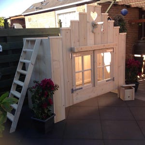 Op de afbeelding: Een houten speelhuisje met een hartvormig raam en een ladder. Het speelhuisje is geschilderd in een lichte houtkleur en heeft een klein dak. Het speelhuisje staat op een terras.