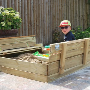 May include: A wooden sandbox with a hinged lid that can be used as a bench. The sandbox is made of natural wood and has a light brown finish. A child is sitting in the sandbox, wearing a yellow hat and sunglasses.
