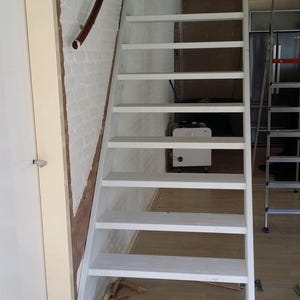 May include: White wooden staircase with a handrail leading up to an attic or loft. The stairs are in a room with a white brick wall and a white door.