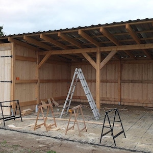 Puede incluir: Un cobertizo de madera con techo de metal y una puerta parcialmente abierta. El cobertizo está en construcción y tiene una escalera y caballetes en el interior. Un coche está aparcado fuera del cobertizo con el maletero abierto.