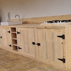 Puede incluir: Una cocina exterior de madera con un fregadero, una estufa de gas y varios armarios. Los armarios tienen bisagras negras y están hechos de madera clara.