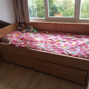 May include: A wooden bed frame with a pink and yellow patterned bedspread. The bed has a pull-out drawer for storage.