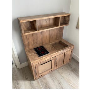 May include: A wooden play kitchen with a black stovetop, two doors, and a shelf above. The kitchen is made of reclaimed wood and has a rustic look.