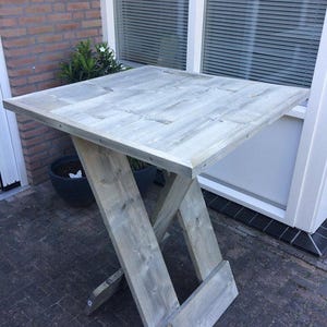 Puede incluir: Una mesa plegable de madera gris con un acabado desgastado. La mesa tiene una tapa cuadrada y una base simple en forma de X.