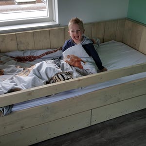 May include: A light wood bed frame with a white mattress and a child lying in it. The bed has a built-in safety rail and a drawer underneath.