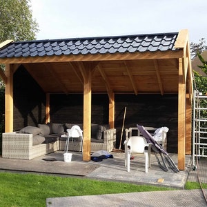 Pode incluir: Uma pérgola de madeira com um telhado de telhas pretas, que fornece sombra sobre uma área de pátio. O pátio é mobiliado com um sofá e cadeiras de vime, e há várias ferramentas e suprimentos espalhados.