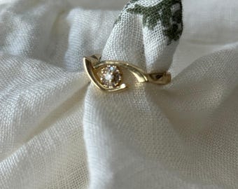 May include: A gold-colored ring with a central, round, clear stone. The ring's design features a curved band that wraps around the finger, with the stone set in a bezel setting. The ring is displayed on a white cloth.
