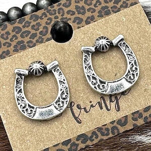 May include: Silver-toned horseshoe earrings with intricate scrollwork and a beaded accent. The earrings are displayed on a brown card with a leopard print border, with the word "Fring" visible. The background includes silver-colored beads.