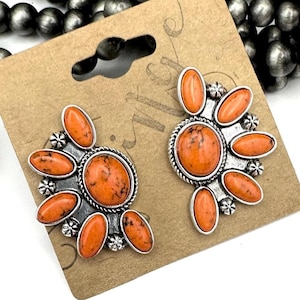 May include: A pair of orange and silver earrings with a southwestern design. The earrings feature a large orange stone surrounded by smaller orange stones.