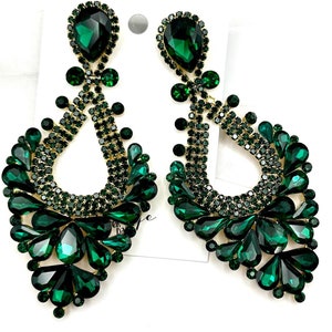 May include: A pair of emerald green chandelier earrings. The earrings feature a teardrop shape with a gold-tone frame and are adorned with numerous green rhinestones. The design includes large, faceted stones and smaller, sparkling accents, creating a glamorous look.