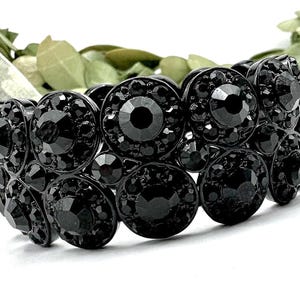 May include: A black stretch bracelet featuring multiple rows of large, round, black faceted beads. The beads have a central, smaller, faceted stone. The bracelet is set against a blurred background of greenery.