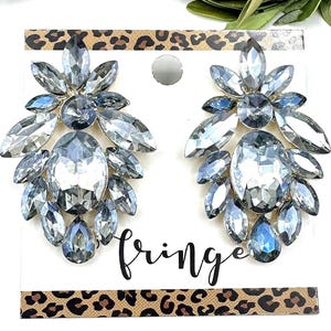 May include: A pair of statement earrings with a cluster of clear and blue-grey faceted crystals. The earrings are on a white card with the word "fringe" in cursive and a leopard print border.