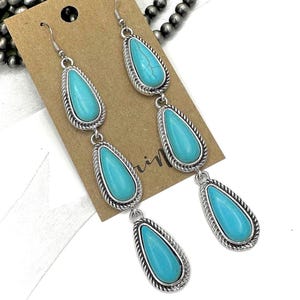 May include: Long, dangling earrings with three turquoise teardrop-shaped stones set in a silver-colored metal frame. The frame has a twisted rope design around the edges. The earrings are displayed on a brown card.