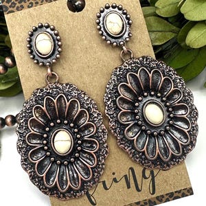 May include: A pair of copper-toned metal drop earrings featuring an oval-shaped floral design with a central white stone. Each earring has a smaller matching oval stud top with a white stone, all mounted on a brown display card with leopard print.