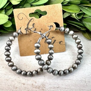 May include: A pair of silver hoop earrings made of small, round beads. The earrings are hanging from a brown card with the word "Boutique" written on it.