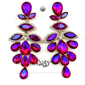 May include: A pair of dangle earrings with a gold base and multiple teardrop-shaped crystals. The crystals are a vibrant iridescent pink and purple color. The earrings are labeled "Bring After Dark".