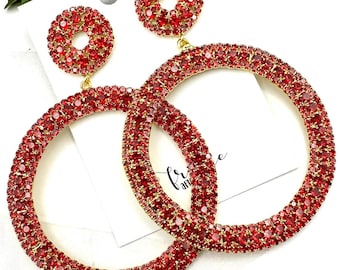 May include: A pair of large hoop earrings with a smaller circle charm dangling from each hoop. The hoops and charms are covered in red rhinestones.