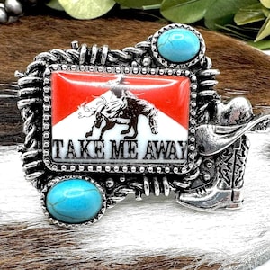 May include: Silver-toned metal belt buckle with a rectangular center featuring a cowboy riding a bull against a red and white background. The buckle has two turquoise stones and the text "TAKE ME AWAY". A cowboy boot and hat are on the right side.