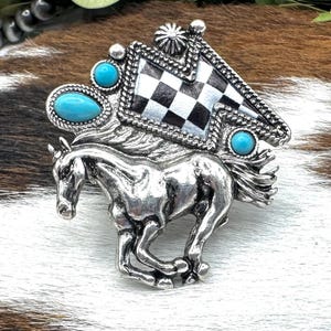 May include: Silver-toned horse-shaped brooch with turquoise accents and a checkered flag design. The brooch features a running horse, oval and round turquoise stones, and a starburst detail. A Western-themed accessory.